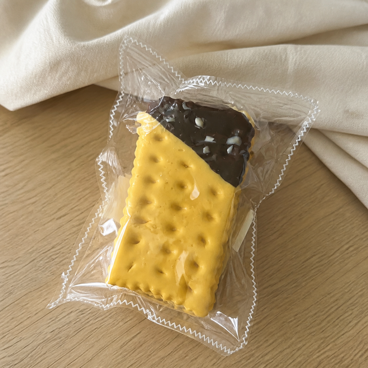 Real product photo of the NOLLUMI Chocolate Butter Sandwich Cookie Squishy in its clear sealed wrapper on a warm neutral setting